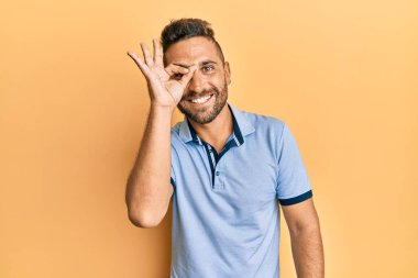 Sakallı, sıradan giyinen yakışıklı bir adam. El hareketleriyle gülümsüyor, gözleriyle mutlu yüzüyle parmaklarının arasından bakıyor.. 