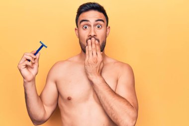 Sakalsız, sakalsız, sarı arka planı jiletle kaplayan, ağzı elle kaplanmış, şok olmuş ve hata yapmaktan korkan genç yakışıklı bir adam. Şaşırtıcı ifade.