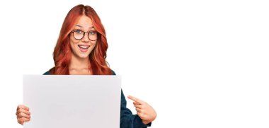 Boş afiş tutan genç kızıl saçlı kadın mutlu bir şekilde el ve parmak işareti yapıyor. 