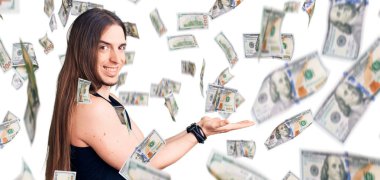 Uzun saçlı, gotik tarzda giyinmiş, siyah elbiseli, avuç içi açık, fotokopi alanını gösteren, mutlu bir gülümseme reklamı sunan genç bir adam.