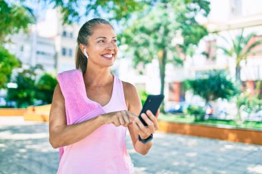 Parkta akıllı telefon kullanarak gülümseyen orta yaşlı sporcu kadın.
