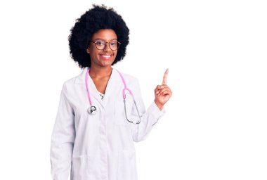 Genç Afro-Amerikalı kadın doktor önlüğü giyiyor ve steteskop takıyor. Yüzünde kocaman bir gülümseme var. Elleriyle kameraya bakıyor.. 