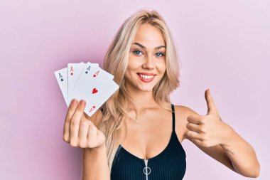 Güzel beyaz sarışın kız elinde as poker kartı tutarken mutlu ve pozitif gülümsüyor, başparmağıyla mükemmel ve onay işareti yapıyor. 