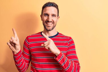 Sıradan çizgili tişört giyen yakışıklı genç adam izole edilmiş sarı arka planda duruyor gülümsüyor ve kameraya bakıyor iki eliyle ve parmaklarıyla yan tarafı işaret ediyor..