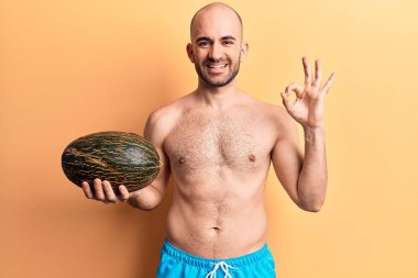 Genç, kel, mayo giyen, elinde kavun tutan, parmaklarıyla imza atan, gülümseyen mükemmel bir sembol olan yakışıklı bir adam. 