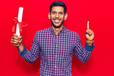 Diploması olan genç Latin bir adam bir fikirle gülümsüyor ya da mutlu bir yüzle işaret ediyor. 