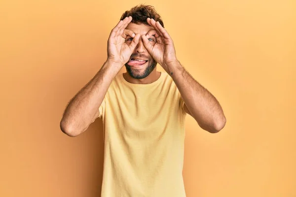 ひげを生やしたハンサムな男は 黄色の背景にカジュアルな黄色のTシャツを着て 双眼鏡が舌を突き出すようなOkジェスチャーをして 目は指を通して見ている クレイジーな表現 — ストック写真