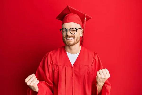 赤い卒業キャップと式のローブを身に着けている若い赤毛の男は非常に満足し 腕を上げ 笑顔と成功のために叫んで勝者のジェスチャーを行うことに興奮しています お祝いのコンセプト — ストック写真