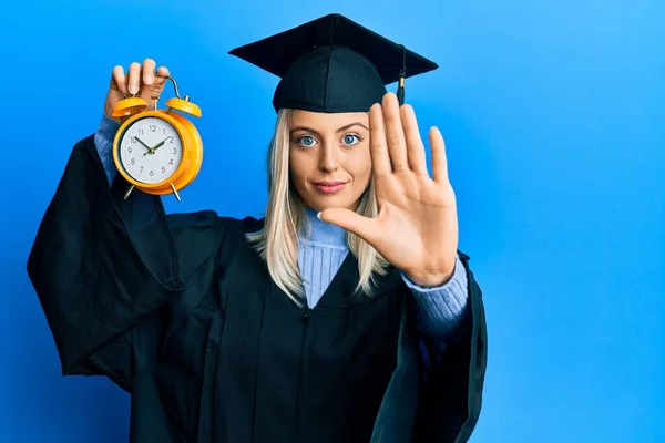 卒業キャップと式のローブを身に着けている美しいブロンドの女性は 目覚まし時計を保持します — ストック写真