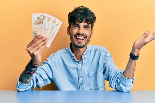 年轻的他的惊慌失措的人手里拿着10英镑的英国钞票坐在桌子上 高兴地微笑着庆祝胜利 高举双手庆祝胜利 — 图库照片