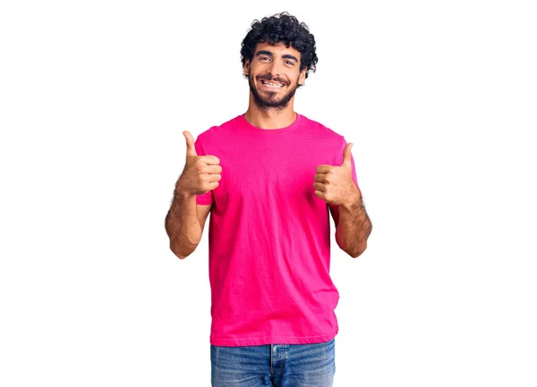 巻き毛のハンサムな若い男と手で積極的なジェスチャーを行うカジュアルなピンクのTシャツの成功サインを身に着けてクマ 親指を笑顔と幸せ 陽気な表情と勝者のジェスチャー — ストック写真