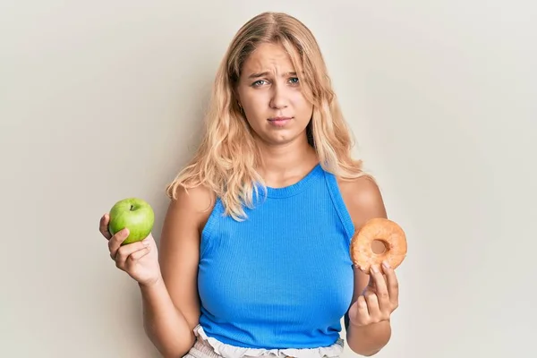 年轻的金发姑娘手里拿着青苹果和甜甜圈 疑神疑鬼 神经质 皱着眉头因为问题而心烦意乱 消极的人 — 图库照片