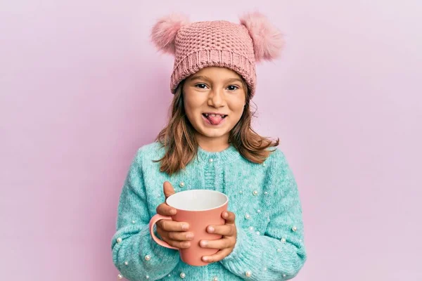 可愛いです女の子保持ピンクマグカップ身に着けています可愛いですウール帽子ステッキング舌アウト幸せなで面白い式 — ストック写真