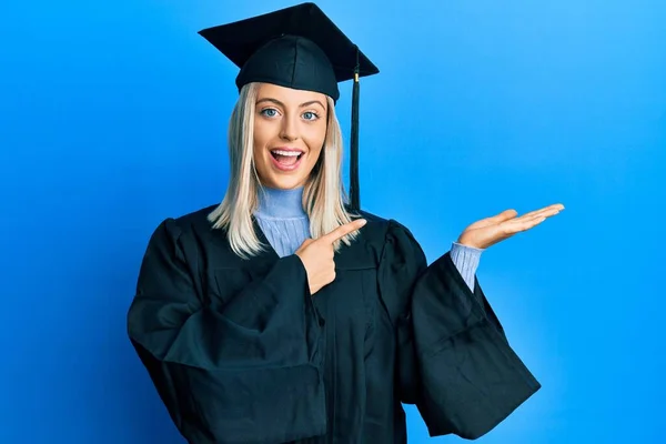 卒業キャップと式のローブを身に着けている美しいブロンドの女性は驚いて 手で提示しながら 指で指してカメラに笑みを浮かべて — ストック写真