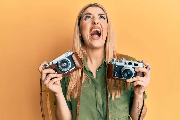 Young caucasian woman holding vintage camera angry and mad screaming frustrated and furious, shouting with anger looking up.