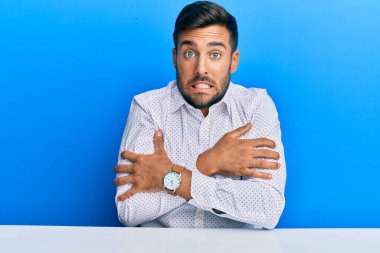 Yakışıklı İspanyol iş kıyafetleri giymiş masada oturmuş soğuktan titriyor ve donuyor yüzünde üzgün ve şok edici bir ifade var. 