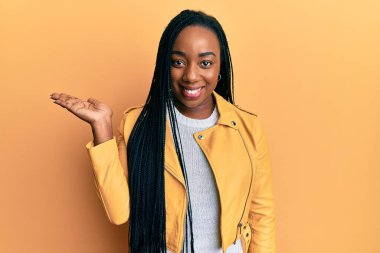 Genç Afrikalı Amerikalı kadın rahat bir ceket giyiyor neşeli bir şekilde gülümsüyor ve avuç içiyle kameraya bakıyor.. 