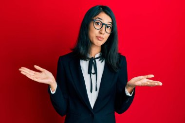 Güzel Asyalı genç bir kadın iş elbisesi giyiyor ve elleri ve kolları havada, kafası karışmış bir ifade takınıyor. şüphe kavramı. 