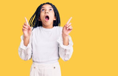 Beyaz bir tişört giyen tatlı Afro-Amerikalı kız şaşırmış ve şaşırmış bir şekilde yukarı bakıyor ve parmaklarıyla işaret ediyor ve kollarını kaldırıyor.. 