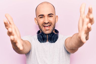 Genç yakışıklı kel adam müzik dinliyor kulaklıkla kameraya bakıyor kucaklaşmak için kollarını açmış gülümsüyor. Mutluluğu kucaklayan neşeli bir ifade.. 