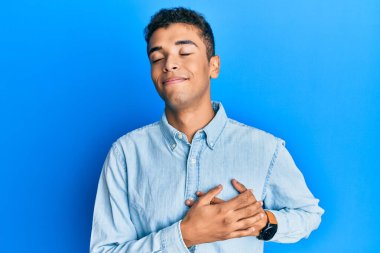 Genç, yakışıklı, Afro-Amerikalı sıradan kıyafetler giyen, elleri göğsünde gülümseyen, gözleri kapalı, yüzünde minnet dolu bir jest olan bir adam. sağlık kavramı. 