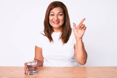 Orta yaşlı esmer kadın masaya oturmuş bir bardak su içen mutlu bir şekilde elini ve parmağını kenarı işaret ederek gülümsüyor. 