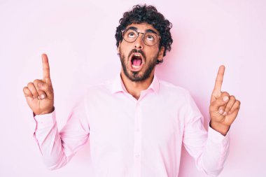 Kıvırcık saçlı ve ayı gibi iş gömleği ve gözlüklü yakışıklı genç adam şaşırmış ve şaşırmış bir şekilde yukarı bakıyor ve parmaklarıyla işaret ediyor ve kollarını kaldırıyor.. 