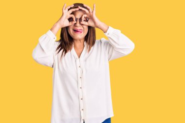 Orta yaşlı Latin kadın, günlük kıyafetler ve gözlük takıyor dürbün gibi jest yapıyor, dili dışarı çıkarıyor, gözleri parmaklarından bakıyor. çılgın ifade. 