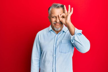 Orta yaşlı, gri saçlı, günlük kıyafetler giyen, el hareketleriyle gülümseyen, mutlu yüzüyle parmaklarının arasından bakan bir adam.. 