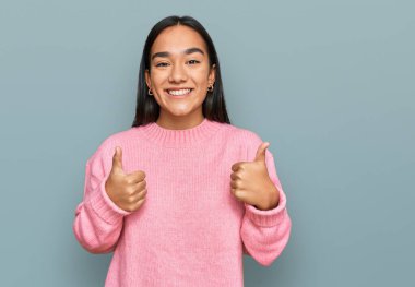 Asyalı genç bir kadın, günlük kışlık süveter giyer, başarı işareti yapar, elleriyle olumlu jest yapar, gülümser ve mutlu olur. Neşeli ifade ve kazanan jest. 