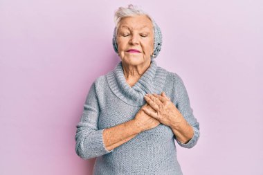 Yaşlı, gri saçlı, yün kazak giyen, kış şapkalı elleri göğsünde, gözleri kapalı gülümseyen ve yüzünde minnet ifadesi olan bir kadın. sağlık kavramı. 