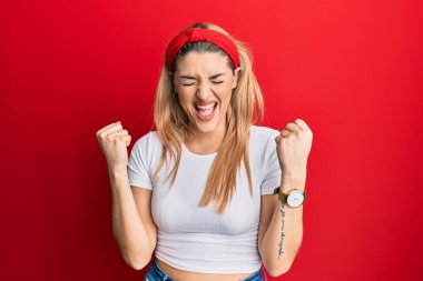 Beyaz gündelik tişört giyen genç beyaz kadın mutlu ve heyecanlı bir şekilde kollarını kaldırıp, gülümseyerek ve başarı için bağırarak kazanan jestler yapıyor. kutlama konsepti. 