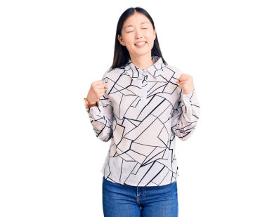 Genç, güzel Çinli bir kadın, gündelik tişört giyip çok mutlu ve heyecanlı bir şekilde kolları havada, gülümseyerek ve başarı için çığlık atarak kazanan bir jest yapıyor. kutlama konsepti. 