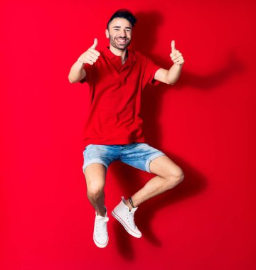 Şapkalı genç teslimatçı mutlu gülümsüyor. Yüzünde tebessümle zıplıyor. Tamam işareti yapıyor. Kırmızı arka plan üzerinde başparmaklar yukarı doğru.