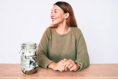 Masada içi dolar dolu bir kavanozla oturan genç esmer kadın yüzünde gülümsemeyle yan yana bakıyor, doğal bir ifade. Kendinden emin gülen. 