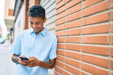 Genç Latin adam mutlu bir şekilde gülümsüyor. Şehrin duvarına yaslanmış akıllı telefon kullanıyor..