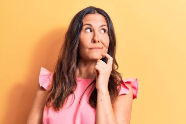 Genç, güzel, esmer, pembe bir tişört giyen, sarı arka planda duran, parmakları çenesinde olan, kuşkuya odaklanmış bir kadın.