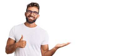 Latin asıllı genç adam günlük kıyafetler ve gözlük takıyor avuç içi gösteriyor ve baş parmaklarıyla iyi bir jest yapıyor, mutlu ve neşeli gülümsüyor. 
