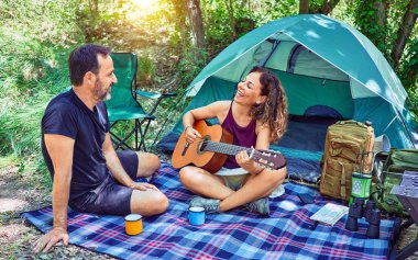 Ormanda kamp yaparken gülümseyen orta yaşlı bir çift. Yerde oturup klasik gitar çalarak şarkı söylemek.
