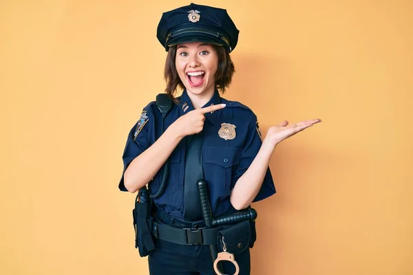 Young beautiful woman wearing police uniform amazed and smiling to the camera while presenting with hand and pointing with finger.