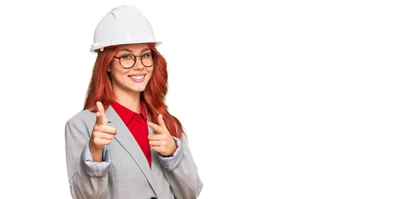 年轻的红头发女人戴着建筑师的硬礼帽 手指指向镜头 脸上洋溢着欢乐和滑稽的表情 充沛的精力和活力 — 图库照片