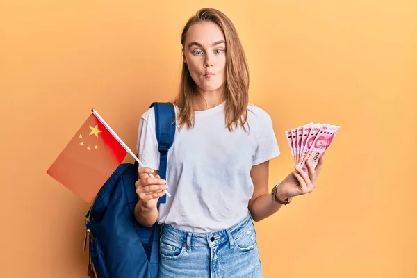 Beautiful blonde woman exchange student holding china flag and yuan banknotes making fish face with mouth and squinting eyes, crazy and comical.