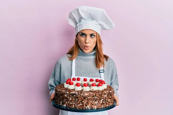 Hispanic young chef woman holding chocolate cake making fish face with mouth and squinting eyes, crazy and comical.
