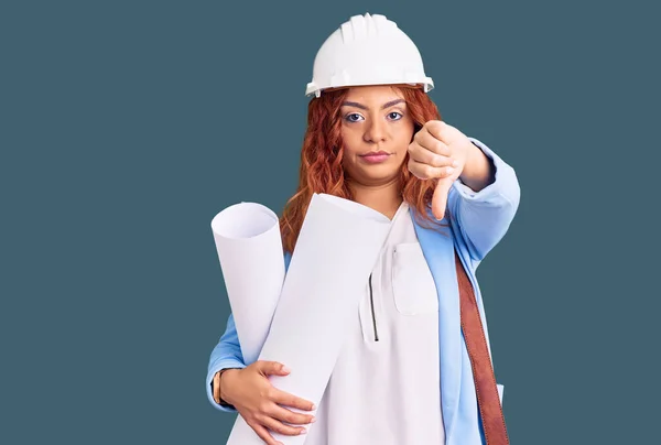 年轻的拉丁女人戴着建筑师的硬礼帽 头戴皮包 手里拿着愤怒的蓝图 脸上挂着不喜欢戴大拇指的负面标志 还有拒绝的念头 — 图库照片