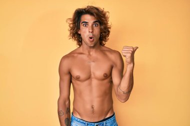 Young hispanic man standing shirtless surprised pointing with hand finger to the side, open mouth amazed expression. 