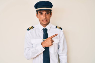 Young hispanic man wearing airplane pilot uniform pointing aside worried and nervous with forefinger, concerned and surprised expression 