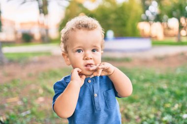 Üzgün küçük çocuk, güneşli bir günde parkta diş etlerine dokunduğu için parmaklarını dudaklarına koyuyor. Güzel sarı saçlı erkek bebek yeni bebek dişleri için acı çekiyor.