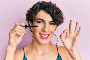 Kirpik rimel tutan genç adam makyaj yapıyor. Parmaklarıyla imza atıyor. Gülümsüyor. 