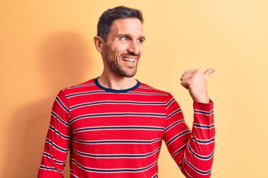 Sıradan çizgili tişört giyen yakışıklı genç adam izole edilmiş sarı arka planda dikiliyor baş parmağını yana kaldırıyor ve ağzı açık gülümsüyor.