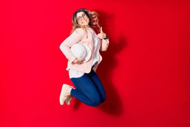 Genç, sarışın, gözlüklü, mimar şapkası takan mutlu bir kadın. Yüzünde tebessümle zıplıyor Tamam işareti yapıyor başparmağınla izole edilmiş kırmızı arka planda